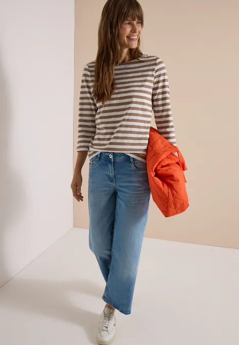 Basic Shirt met boothals en strepen natural taupe Frau in gestreiftem Pullover, weiten Jeans und Sneakern hält eine orange Jacke und lächelt.