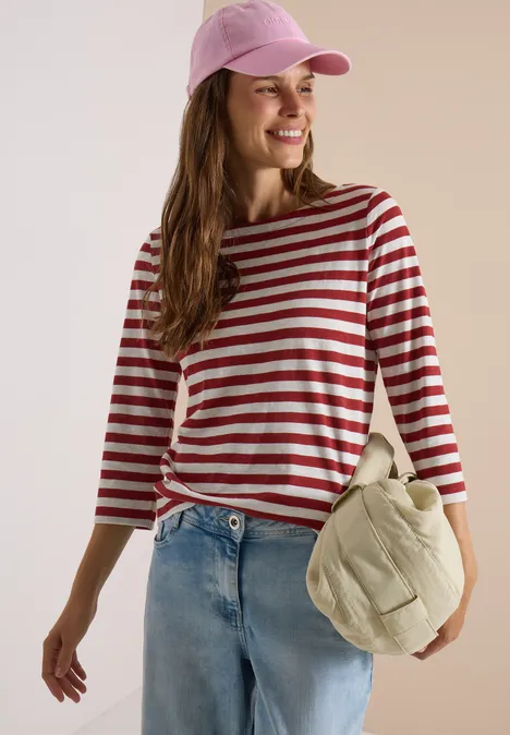Vrouw in rood-wit gestreept shirt, spijkerbroek en roze pet, met een nonchalante tas, glimlachend.