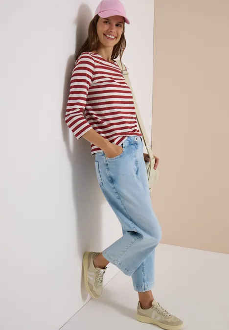 Frau mit rosa Cap, rot-weiß gestreiftem Shirt, lockeren Jeans und Sneakers, lächelnd an eine Wand gelehnt.