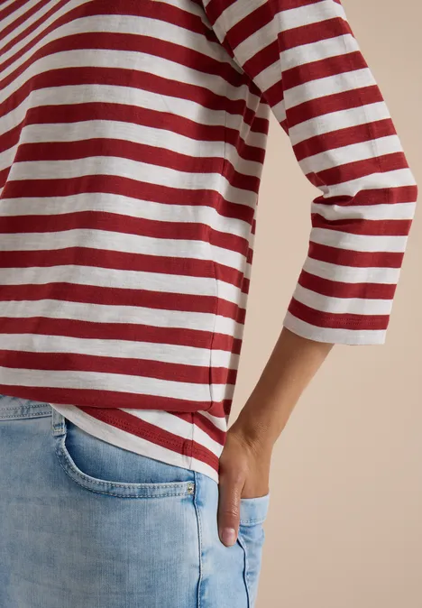 Person trägt rot-weiß gestreiftes 3/4-Arm-Shirt und helle Jeans, eine Hand in der Hosentasche.