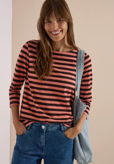 Vrouw lacht in gestreept 3/4-mouwshirt en jeans, handen in zakken, voor neutrale beige achtergrond.
