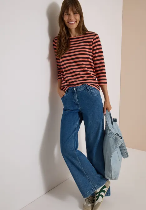Lächelnde Frau lehnt an Wand, trägt gestreiftes Shirt, weite Jeans, Sneaker und hält Jeansjacke.