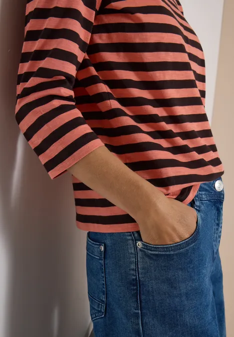 Person lehnt an Wand, trägt rot-schwarz gestreiftes Shirt und blaue Jeans, eine Hand in der Tasche.