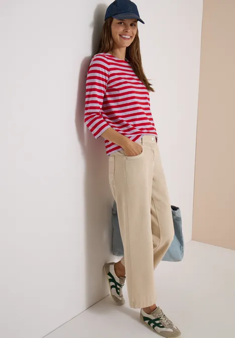 Basic Shirt mit Boat Neck und Streifen signal red Junge Frau im rot-weiß gestreiften Pullover, beige Hose und Cap lehnt lächelnd an Wand und hält Tasche.