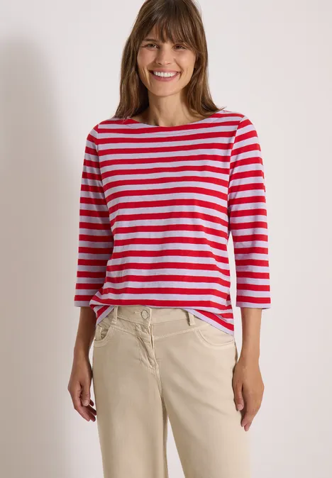 Basic Shirt mit Boat Neck und Streifen signal red Eine Frau in rotem-weiß gestreiftem Dreiviertelarmshirt und beiger Hose lächelt vor hellem Hintergrund.