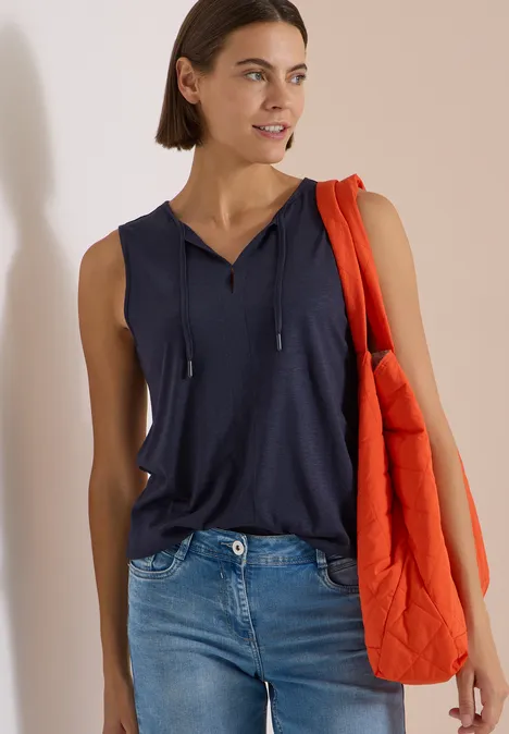 Frau in ärmellosem dunkelblauem Top und Jeans, trägt eine orange Steppjacke über der Schulter.