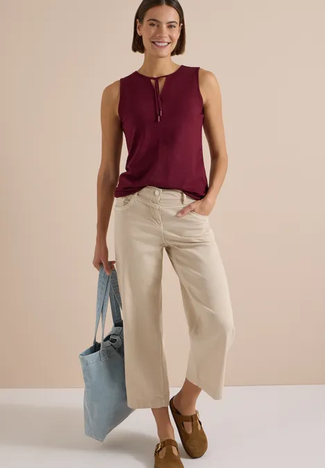 Frau in beigen Culottes, weinrotem ärmellosem Top, braunen Mokassins, blauem Stoffbeutel.