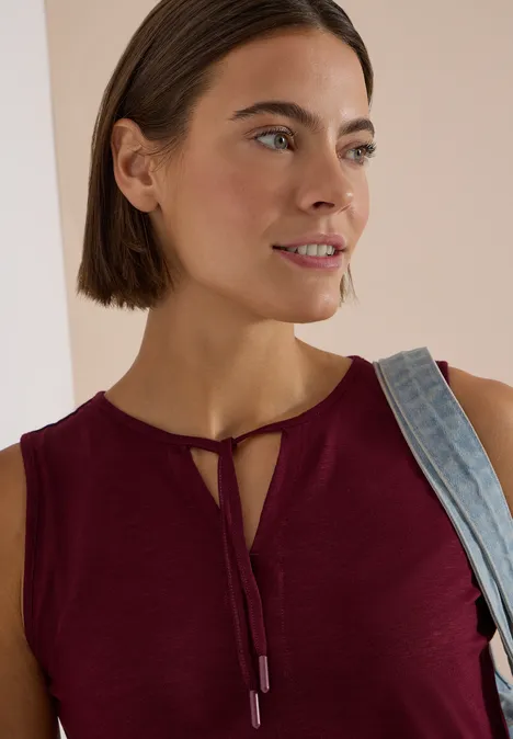Frau mit kurzem braunen Haar in ärmellosem bordeauxfarbenem Top mit Jeans-Träger, lächelnd