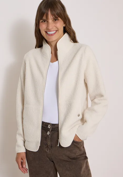 Frau in cremeweißer Fleecejacke, weißem Shirt und braunen Cordhosen, lächelt.