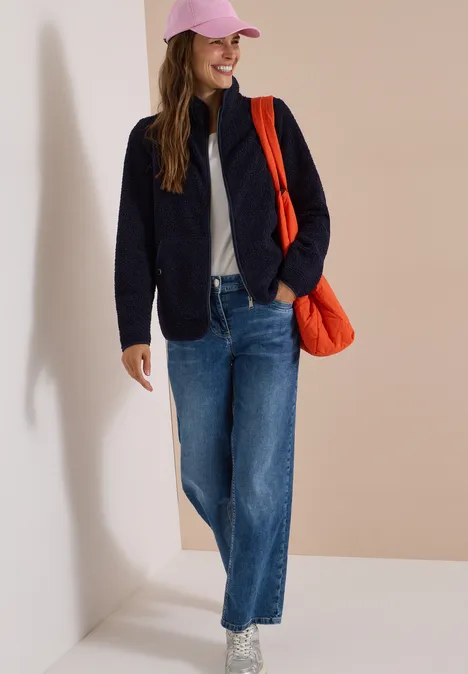 Eine Frau in Jeans, Steppjacke und rosa Kappe trägt eine orangene Tasche und lächelt.