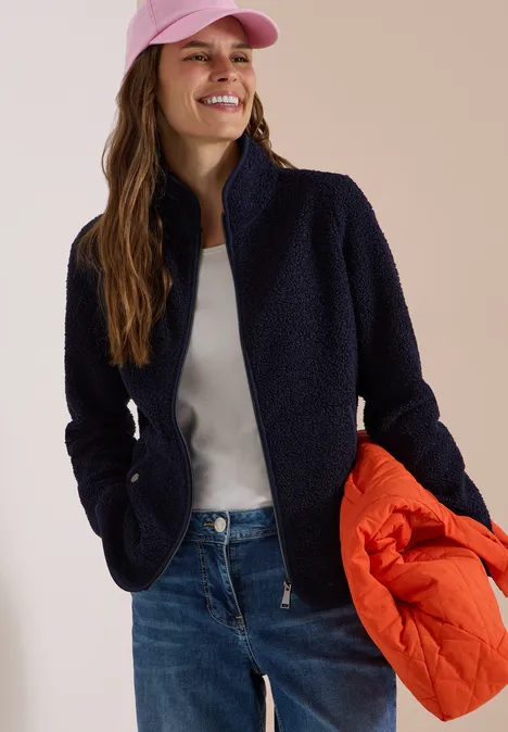 Frau lächelnd in Jeans, weißem Shirt, schwarzem Fleece, rosa Cap, hält orange Steppjacke.