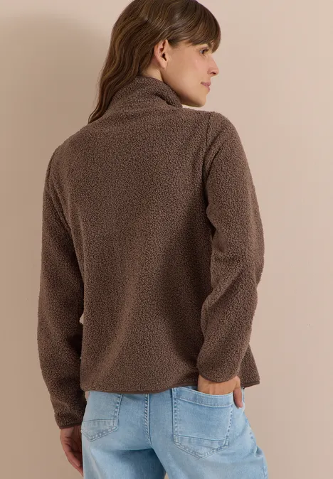 Frau von hinten in brauner Fleecejacke und blauen Jeans vor neutralem Hintergrund.