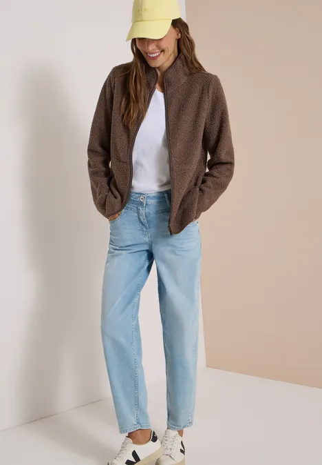 Frau in brauner Jacke, weißem T-Shirt, hellblauen Jeans, weißen Sneakern und beiger Cap.