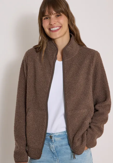 Frau lächelt in brauner Fleecejacke, weißem Shirt und Jeans vor neutralem Hintergrund