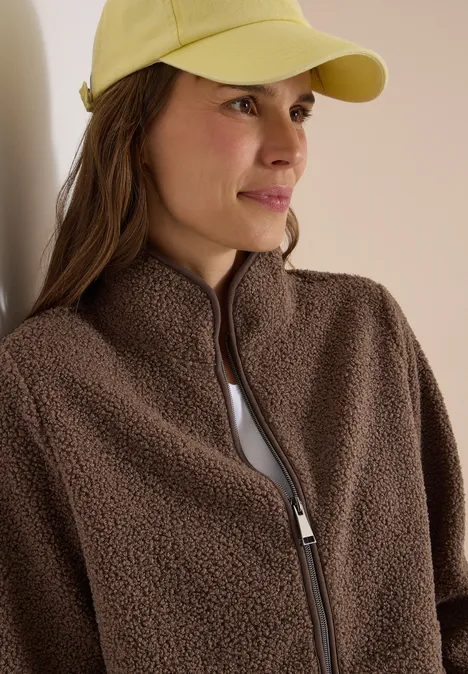 Frau mit gelber Kappe und brauner Fleecejacke, blickt seitlich.