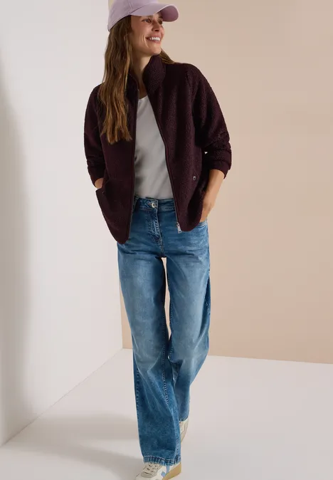 Frau in Jeans, grauem T-Shirt, rosa Cap und dunkelrotem Cardigan steht lässig vor einer Wand.