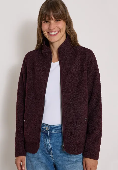 Lächelnde Frau trägt eine violette Strickjacke über weißem T-Shirt und Jeans.