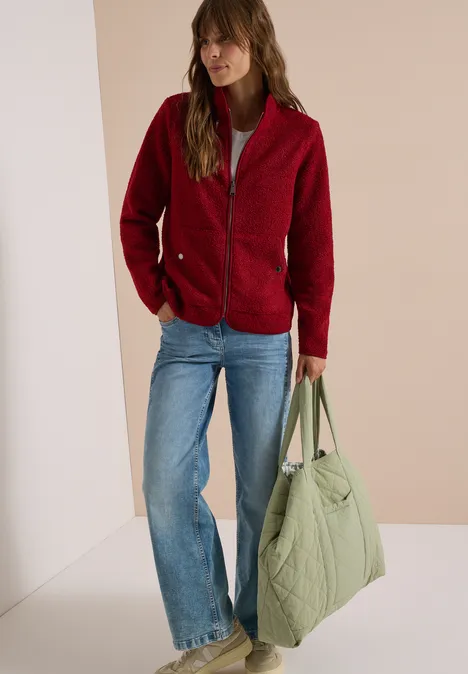 Frau trägt rote Fleecejacke, blaue Jeans und hält große grüne Stepp-Tasche.
