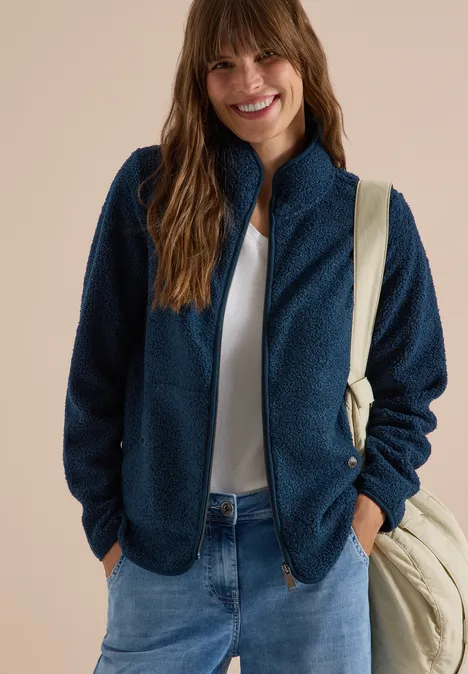 Lächelnde Frau in marineblauer Fleecejacke, weißem T-Shirt, Jeans und Umhängetasche.