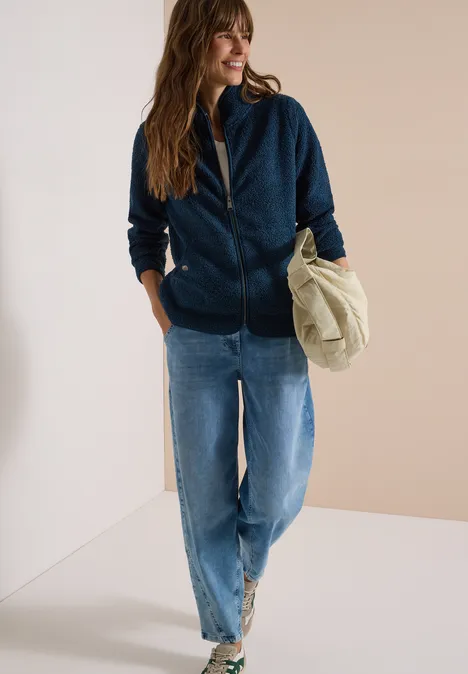 Frau in lässigem Outfit: dunkelblaue Kapuzenjacke, Jeans, weiße Sneaker, hält eine beige Tasche.