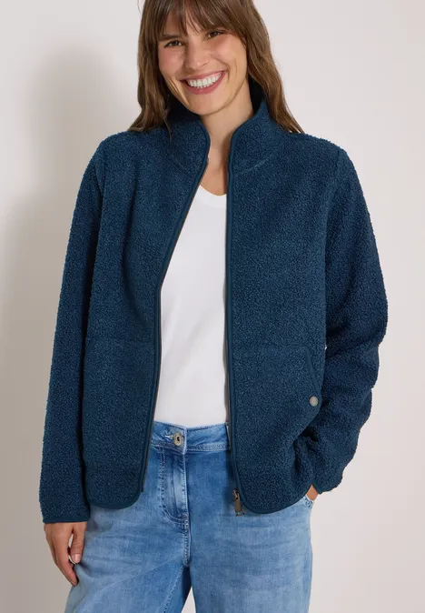 Frau in marineblauer Teddy-Fleecejacke mit Reißverschluss, weißem Shirt und blauen Jeans, lächelnd.