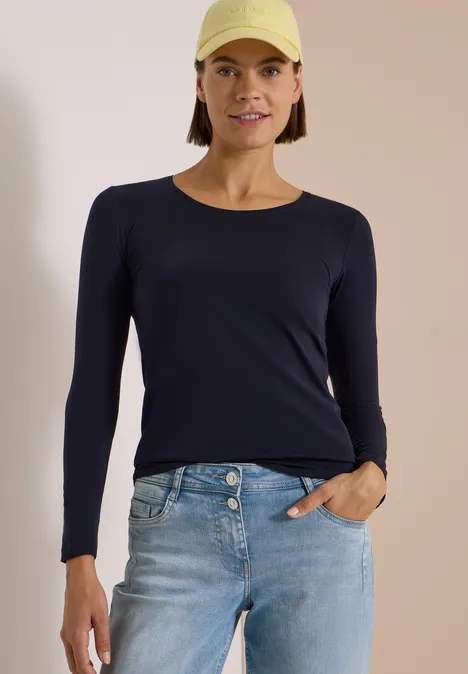 Frau mit gelber Cap, dunkelblauem Langarmshirt und hellen Jeans.