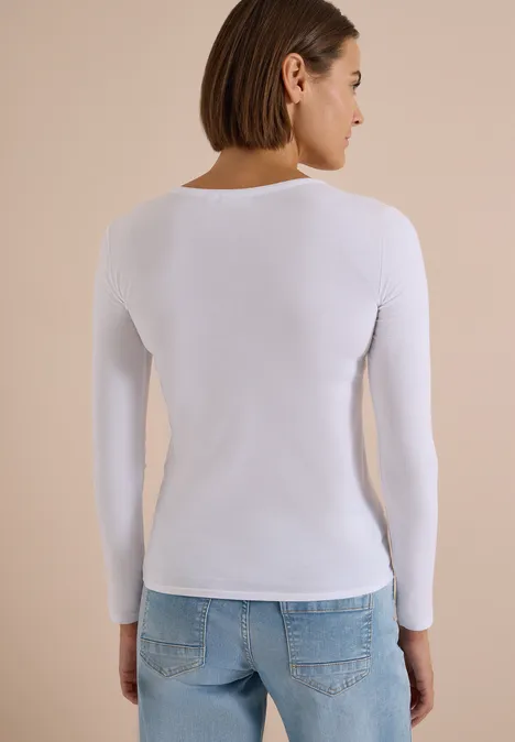 Frau von hinten in weißem Langarmshirt und hellblauen Jeans vor einfarbigem, beigem Hintergrund.