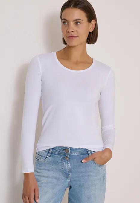 Eine Frau in weißem Langarmshirt und blauen Jeans steht vor neutralem Hintergrund.