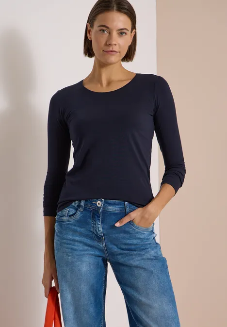 Frau in dunkelblauem 3/4-Shirt und Jeans, mit roter Tasche vor zweifarbigem Hintergrund.