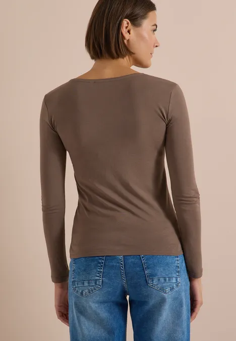 Rückansicht einer Frau mit braunem Langarmshirt und blauen Jeans vor neutralem Hintergrund.
