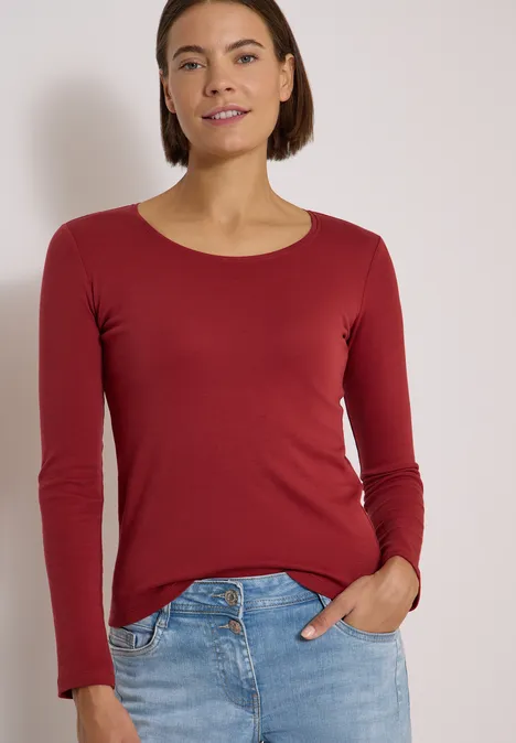 Frau in rotem Langarmshirt und Jeans, lächelnd, eine Hand in der Gesäßtasche.