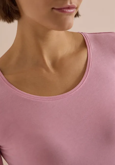 Nahaufnahme des Halses und Oberkörpers einer Person in rosa Oberteil vor hellem Hintergrund.