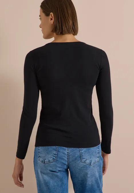 Eine Frau steht mit dem Rücken zur Kamera, trägt ein schwarzes Langarmshirt und blaue Jeans vor beigem Hintergrund.