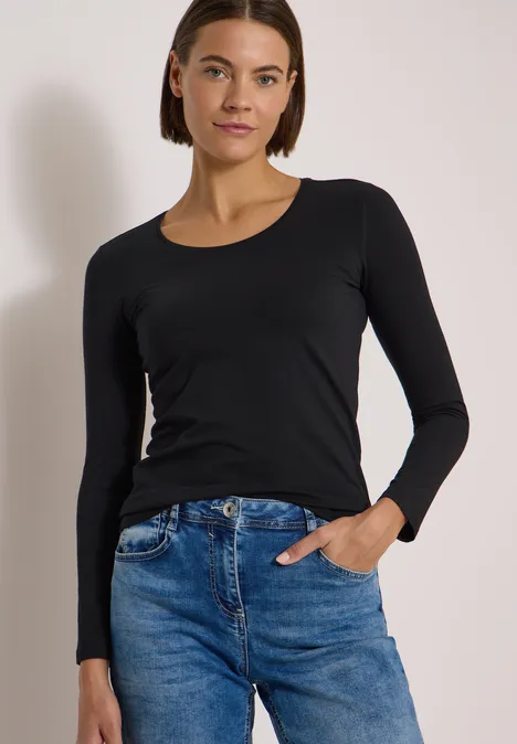 Frau in schwarzem Langarmshirt und blauen Jeans steht vor hellem Hintergrund, lässig mit Hand in Tasche.