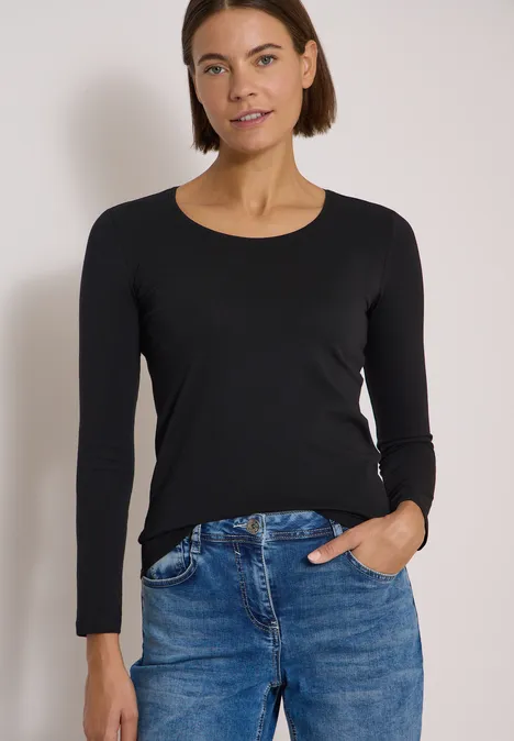 Eine Frau in schwarzem Langarmshirt und blauen Jeans steht locker vor hellem Hintergrund, eine Hand in der Tasche.