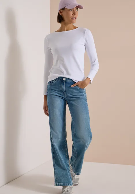 Modell trägt weißes Langarmshirt, blaue Jeans, weiße Sneaker und lila Cap vor schlichter Wand.