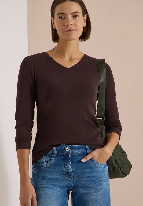 Langarm Shirt in Unifarbe mit V-Neck truffle brown Frau in braunem V-Pullover, blauer Jeans, mit Hand in Tasche und grüner Umhängetasche.