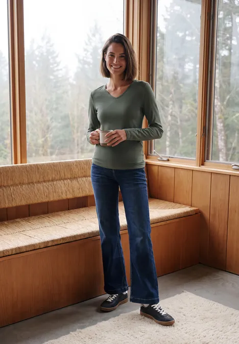 Longsleeve met V-hals dried olive green Frau steht in holzvertäfeltem Raum an großen Fenstern und hält eine Tasse.