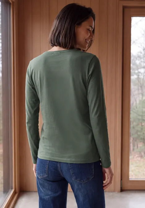 Longsleeve met V-hals dried olive green Frau steht von hinten in grünem Langarmshirt und Jeans vor großen Fenstern.