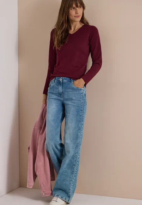 Frau in weiten Jeans und burgunderfarbenem Longsleeve, hält rosa Jacke, vor schlichtem beigem Hintergrund.