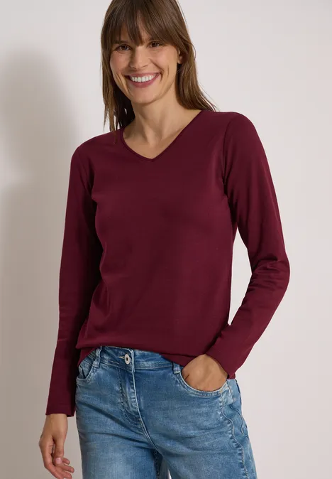 Frau mit langärmligem burgunderfarbenem V-Ausschnittpullover und blauen Jeans, lächelnd vor neutralem Hintergrund.
