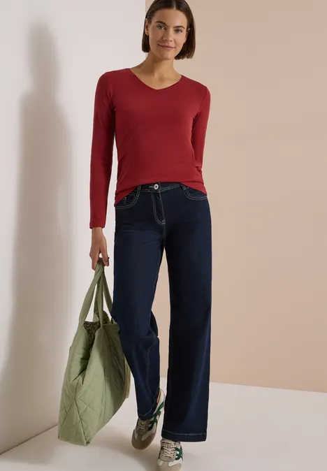Langarm Shirt in Unifarbe mit V-Neck russet red Frau in rotem Langarmshirt, weitem Jeans und grauen Sneakers hält grüne Umhängetasche.