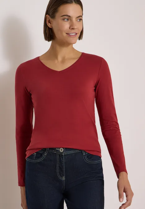 Langarm Shirt in Unifarbe mit V-Neck russet red Frau in rotem Langarmshirt und dunklen Jeans posiert vor hellem Hintergrund.