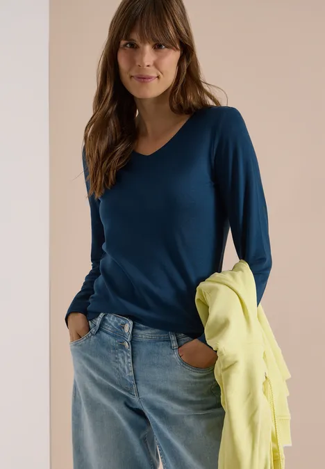Langarm Shirt in Unifarbe mit V-Neck moonlight blue Frau in blauem V-Ausschnitt-Pullover und Jeans hält eine gelbe Jacke und lächelt in die Kamera.
