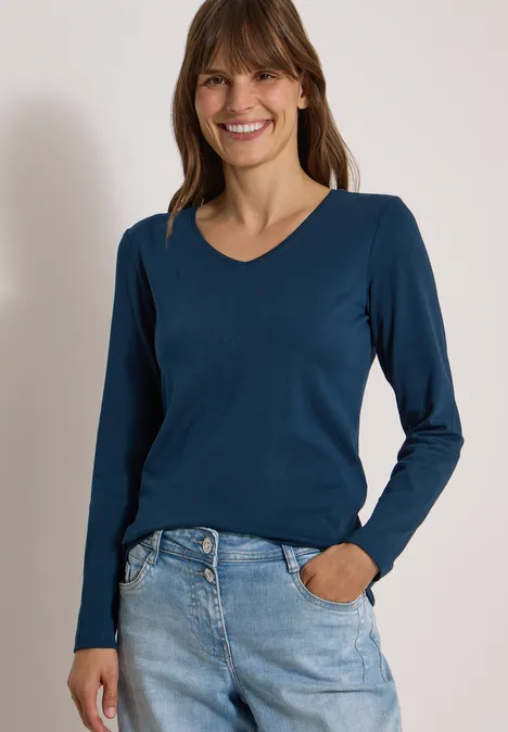 Langarm Shirt in Unifarbe mit V-Neck moonlight blue Frau mit blauem Langarmshirt und Jeans lächelt vor hellem Hintergrund.