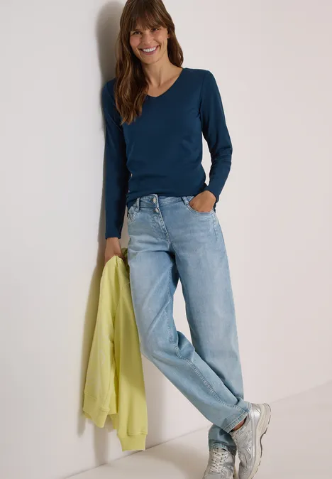Langarm Shirt in Unifarbe mit V-Neck moonlight blue Frau lehnt an Wand, trägt blaues Langarmshirt, helle Jeans und hält einen gelb-grünen Pullover.