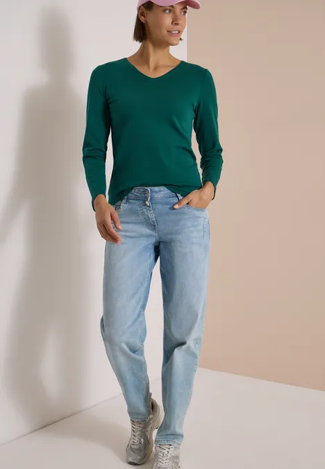 Langarm Shirt in Unifarbe mit V-Neck dark fir green Frau mit grüner V-Pullover, hellblauen Jeans und grauen Sneakern sowie rosa Cap steht vor neutraler Wand.