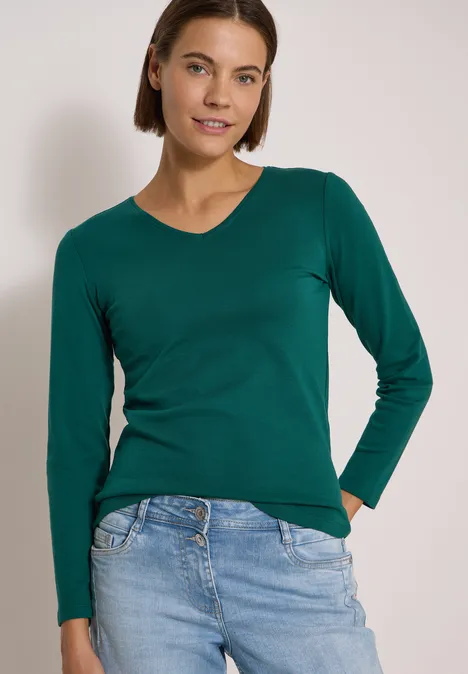 Langarm Shirt in Unifarbe mit V-Neck dark fir green Frau mit kurzem braunem Haar trägt grünes Langarmshirt und helle Jeans, lächelt in die Kamera.