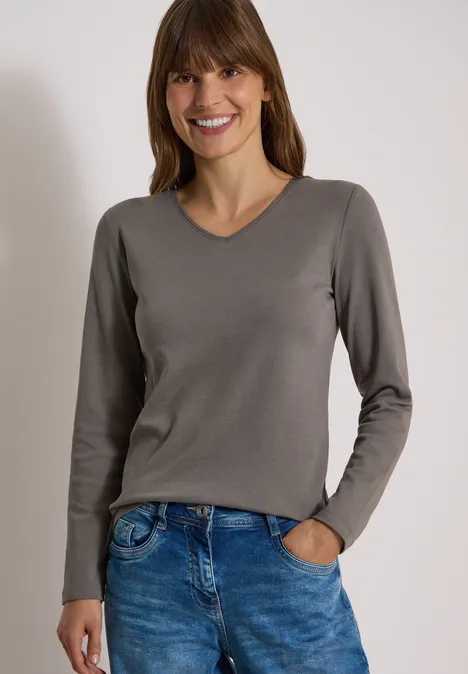 Langarm Shirt in Unifarbe mit V-Neck storm grey Lächelnde Frau in grauem V-Pullover und blauen Jeans, eine Hand lässig in der Hosentasche.