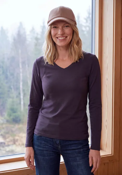 Langarm Shirt in Unifarbe mit V-Neck lilac shadow Eine lächelnde Frau in Jeans, braunem Langarmshirt und Cap vor einem Fenster mit Waldkulisse.